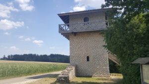 Rekonstruierter Wehrturm aus Stein mit einem umlaufenden Holzgeländer und flachem Dach an einem Feld.