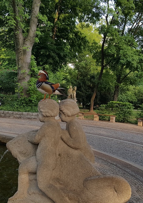 Zwei Kinderfiguren, die auf einem Fisch sitzen und auf dem Kopf der einen Figur sitzt eine Mandarinente.