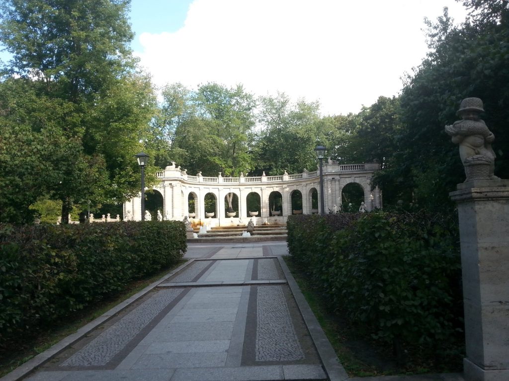 Eingangsbereich zum Märchenbrunnen: gepflasterter Weg mit Blick auf den Brunnen und einen ihn abschließenden Bogen.