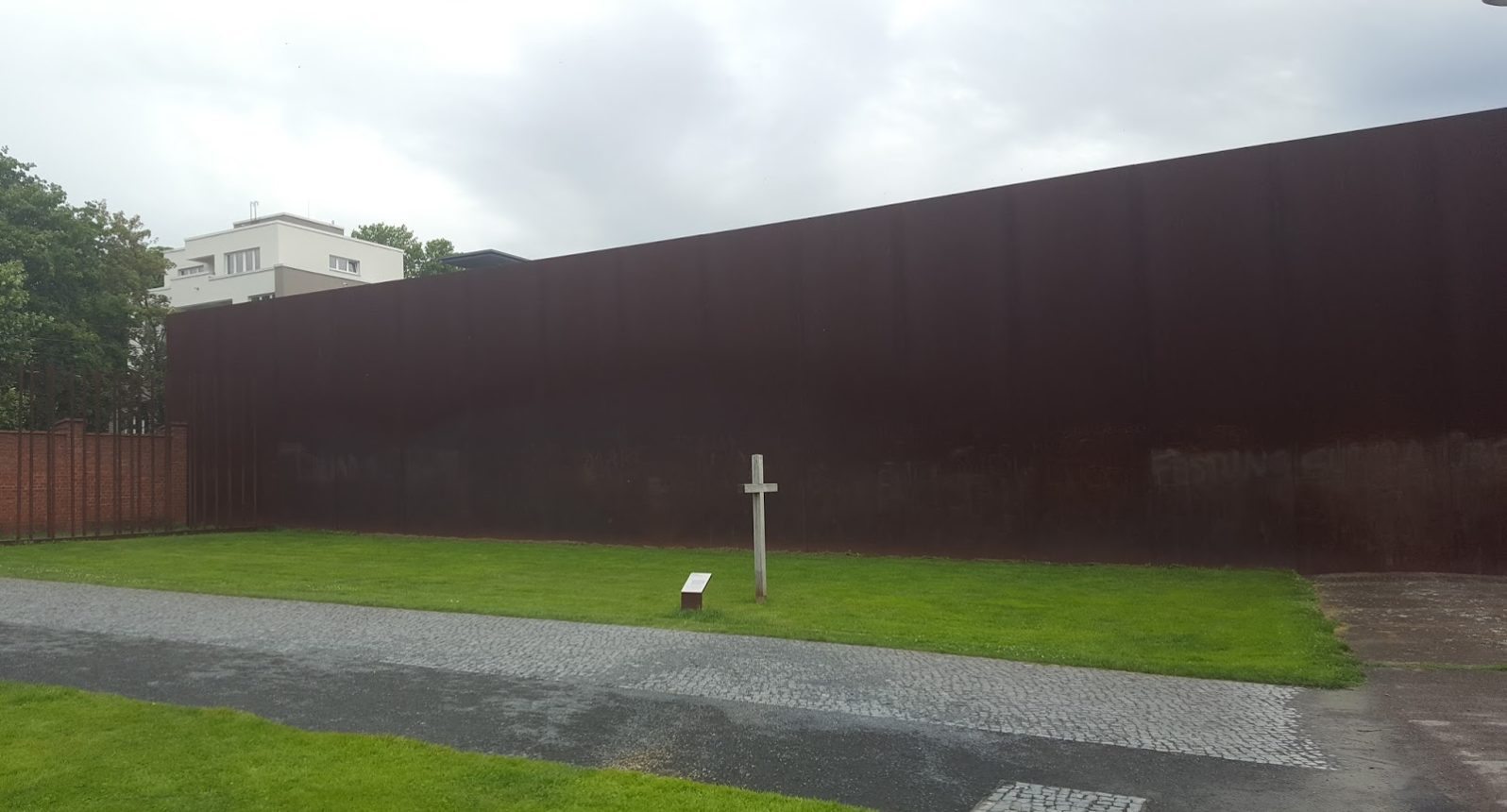 Ein weißes Gedenkkreuz auf grünem Gras vor einer rostbraunen Wand.