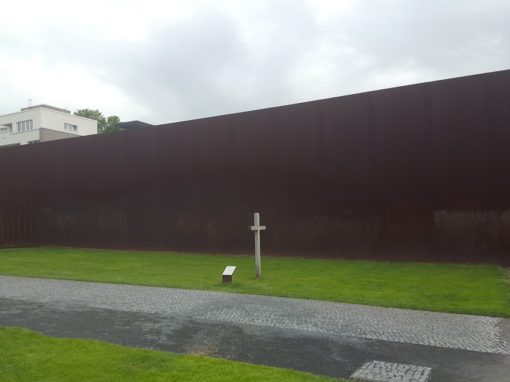 Ein weißes Gedenkkreuz auf grünem Gras vor einer rostbraunen Wand.