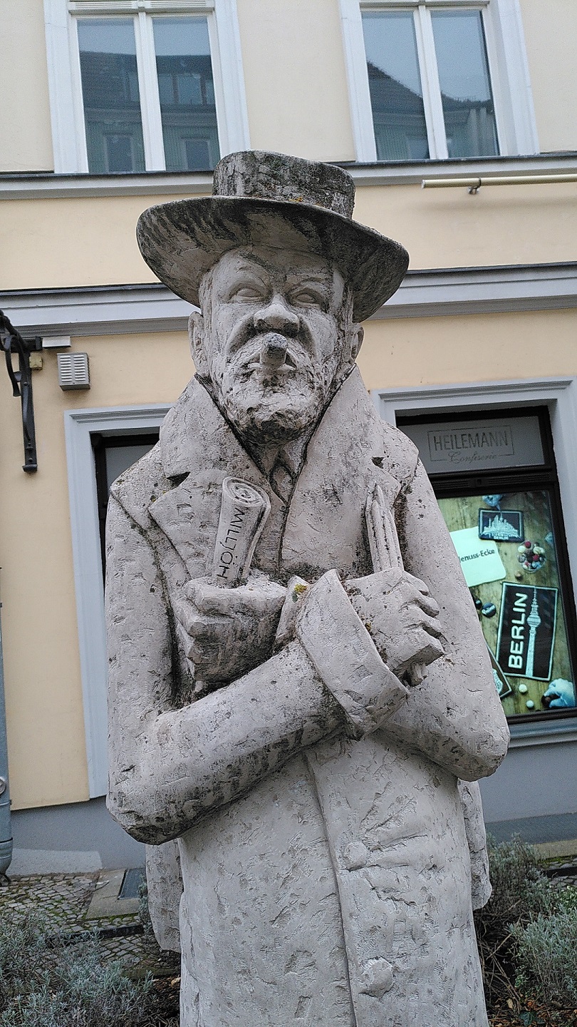 Kalksteinfigur von Zille mit Hut, Zigarre im Mund und ein Blatt Papier in der einen, und Stifte in der anderen Hand.