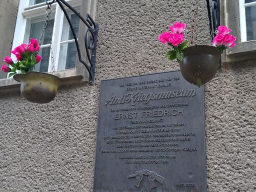 Eine Gedenktafel eingerahmt von zwei Soldatenhelmen, in denen rosa Plastikblumen stecken.