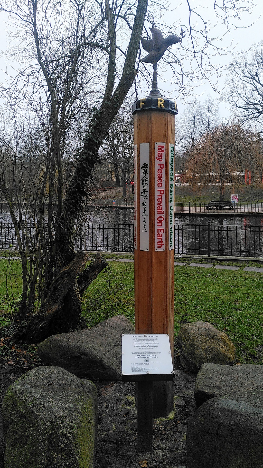 Hölzerner Friedenspfahl mit einer Friedenstaubenfigur auf der Spitze. Im Hintergrund der Teich,