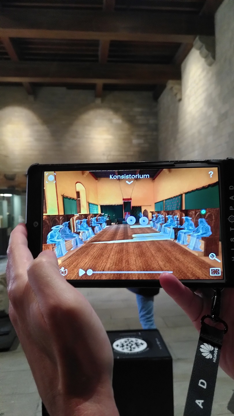 Blick auf das Tablet, das ein animiertes Video zeigt, wie Personen im Konsistorium sitzen. Im Hintergrund nackte Steinmauern.