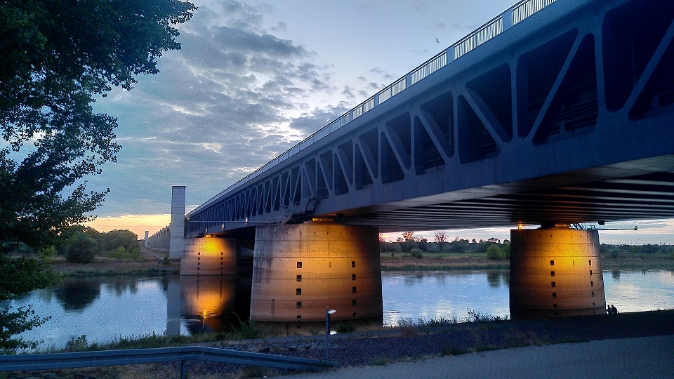 Beleuchtete Kanalbrücke am Abend mit Blick auf den Fluss und im Hintergrund bewölkter Himmel.