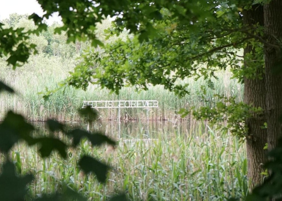Blick durch Bäume auf einen See. Im See ein Schild aus Buchstaben: Art is forever.