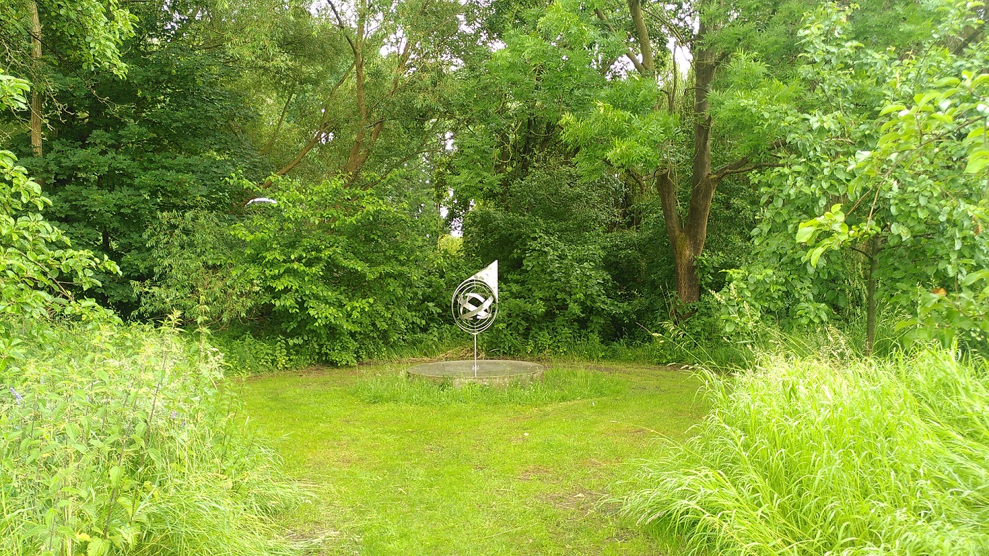 Zwischen hochgewachsenen Gräsern öffnet sich ein gemähter runder Bereich, in dessen Zentrum eine runde Metallskulptur steht. Im Hintergrund Bäume und Büsche.