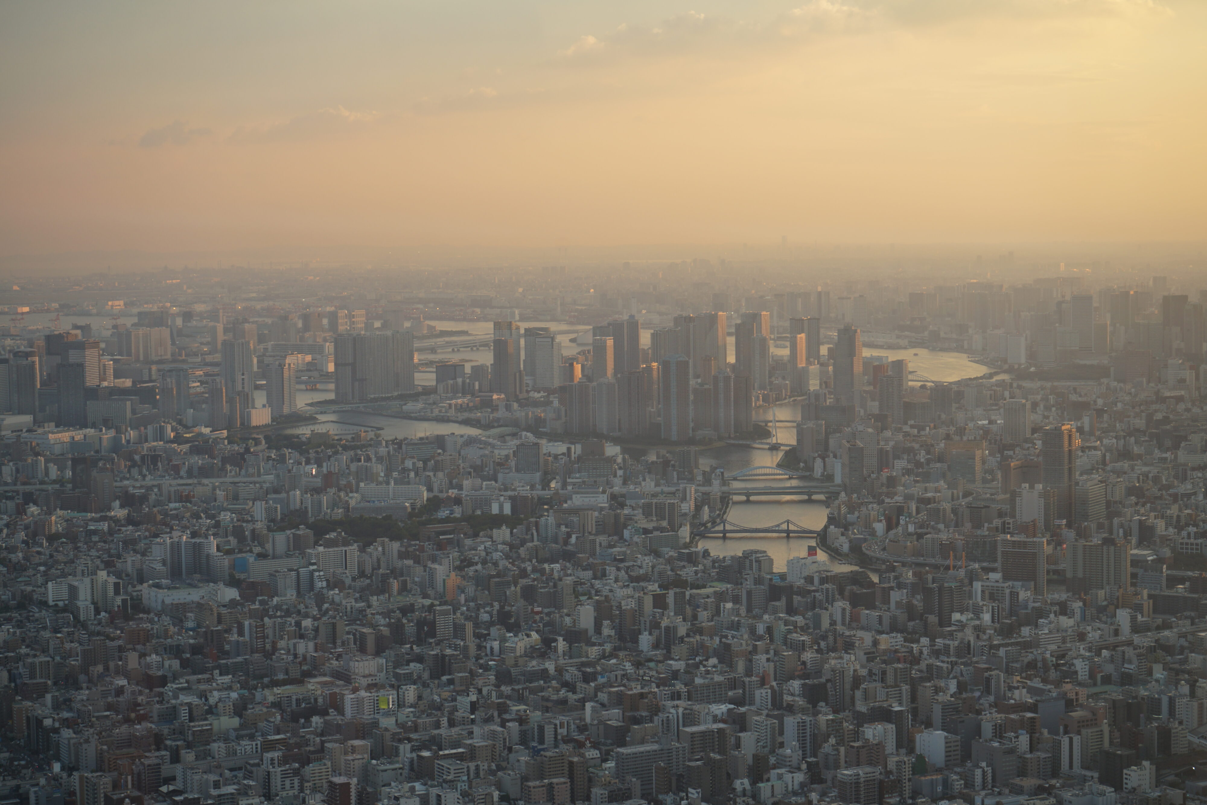 Blick auf Tokyo von oben mit vielen Gebäuden, Fluss und Brücken und einem gelben, diesigen Horizont.