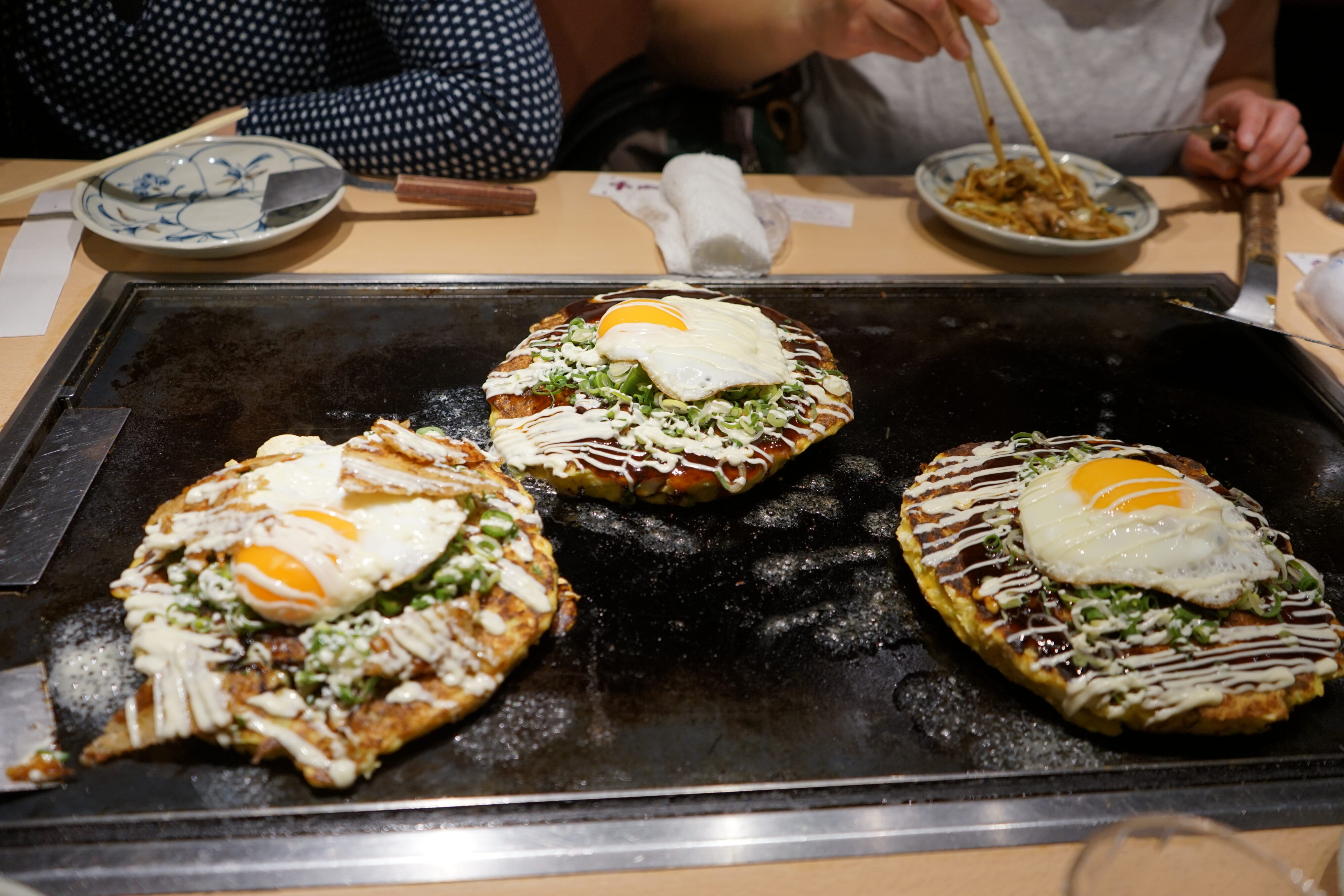 Drei Okonomiyaki mit Spiegelei und Mayonnaise auf einer Wärmefläche.