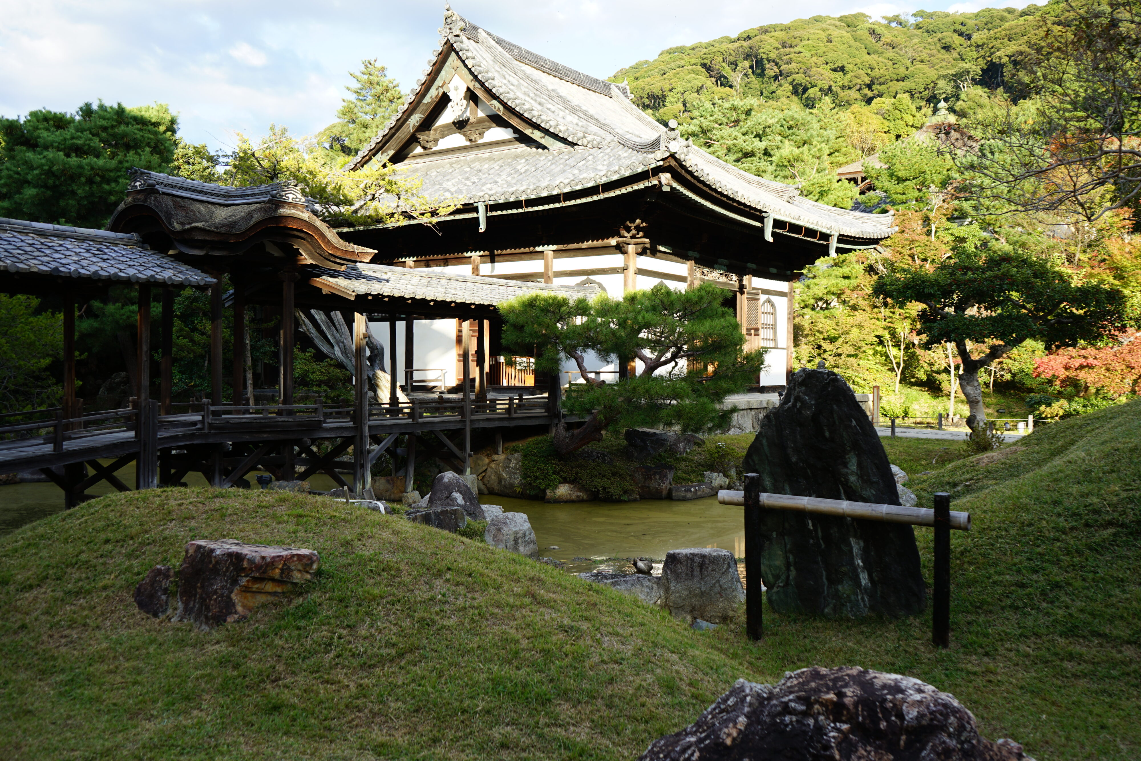 Blick in einen Tempelgarten mit Teich, Steinen, einer Brücke, kleinen Bäumen und einem kleinen Tempelgebäude. Im Hintergrund ein mit Bäumen bewachsener Hügel.