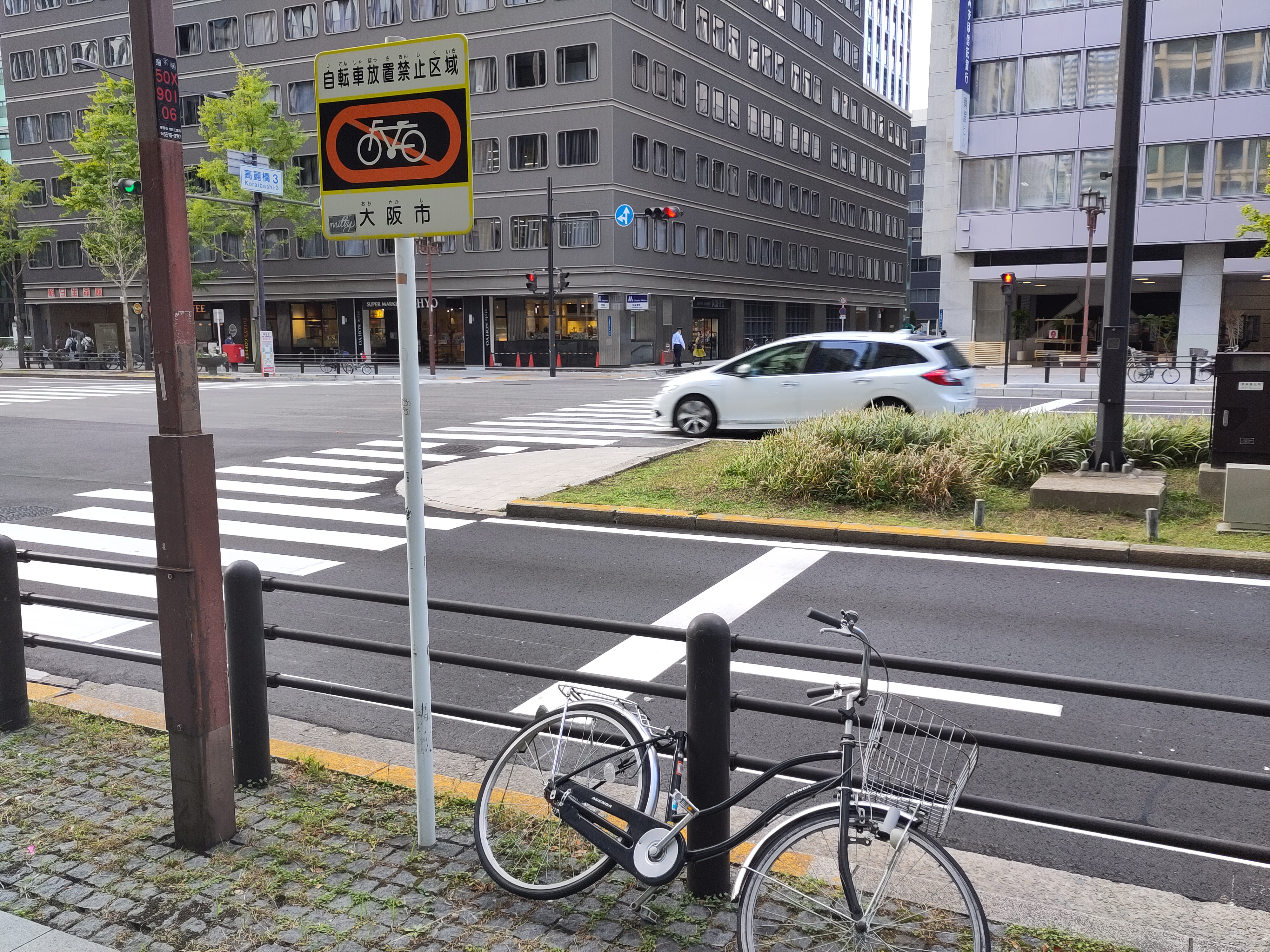 Blick auf eine Straße. Ein Geländer trennt den Bürgersteig von der Straße ab. Ein Schild mit einem durchgestrichenen Fahrrad steht am Geländer, daneben ein angeschlossenes Fahrrad.