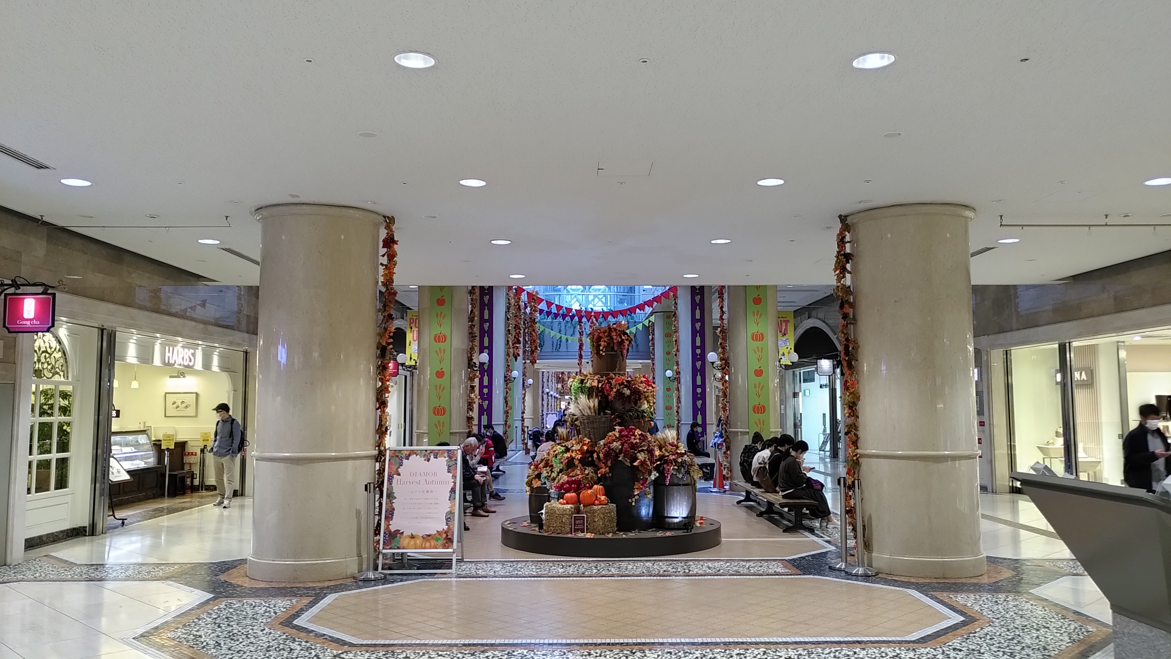 Unterirdische Shoppingmall: im Zentrum ein Arrangement aus Kürbissen und Herbstblumen eingerahmt von Bänken, auf denen Leute sitzen.