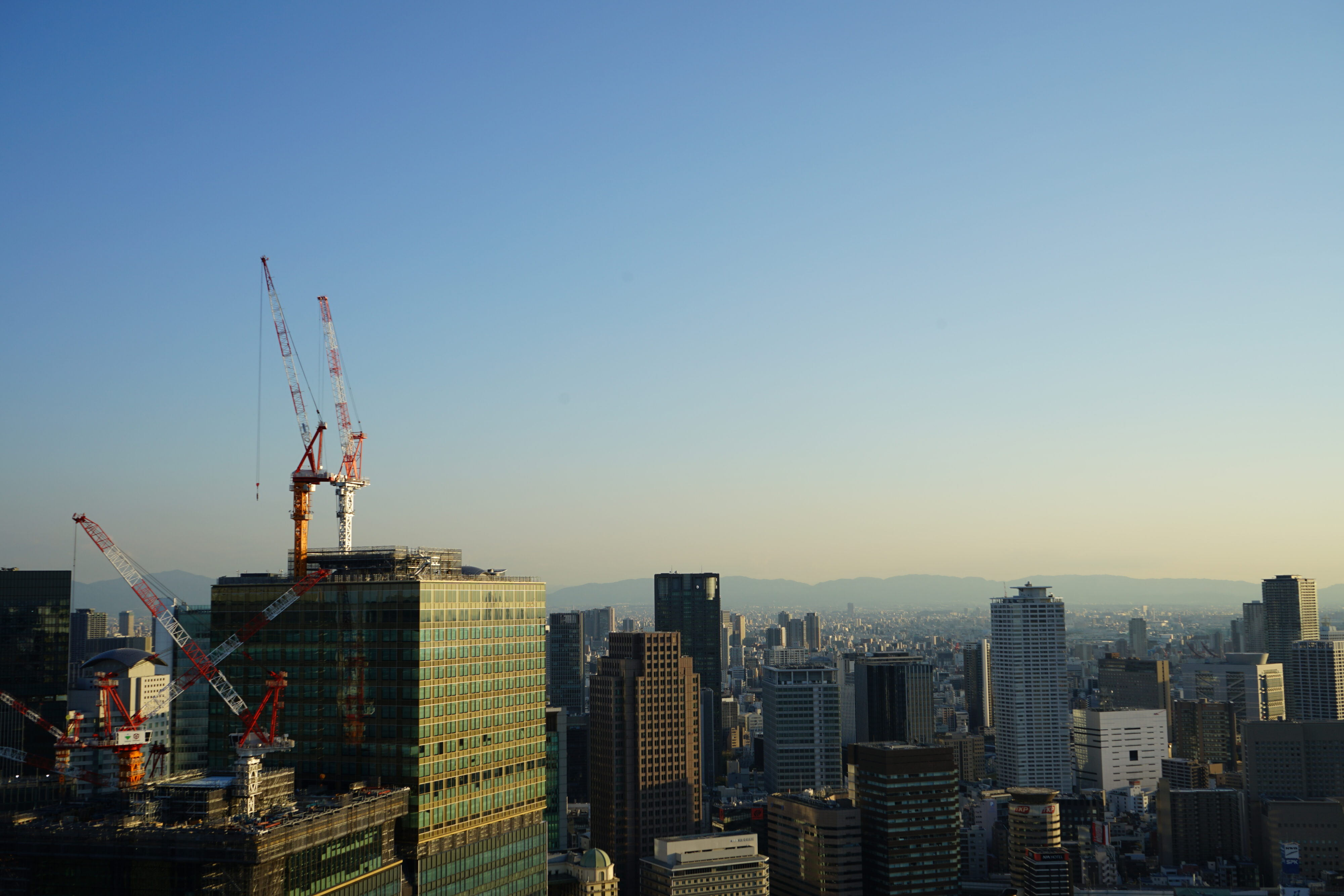 Blick auf Osaka mit Hochhäusern auf den Kräne stehen. In der Ferne sind Berge zu sehen.