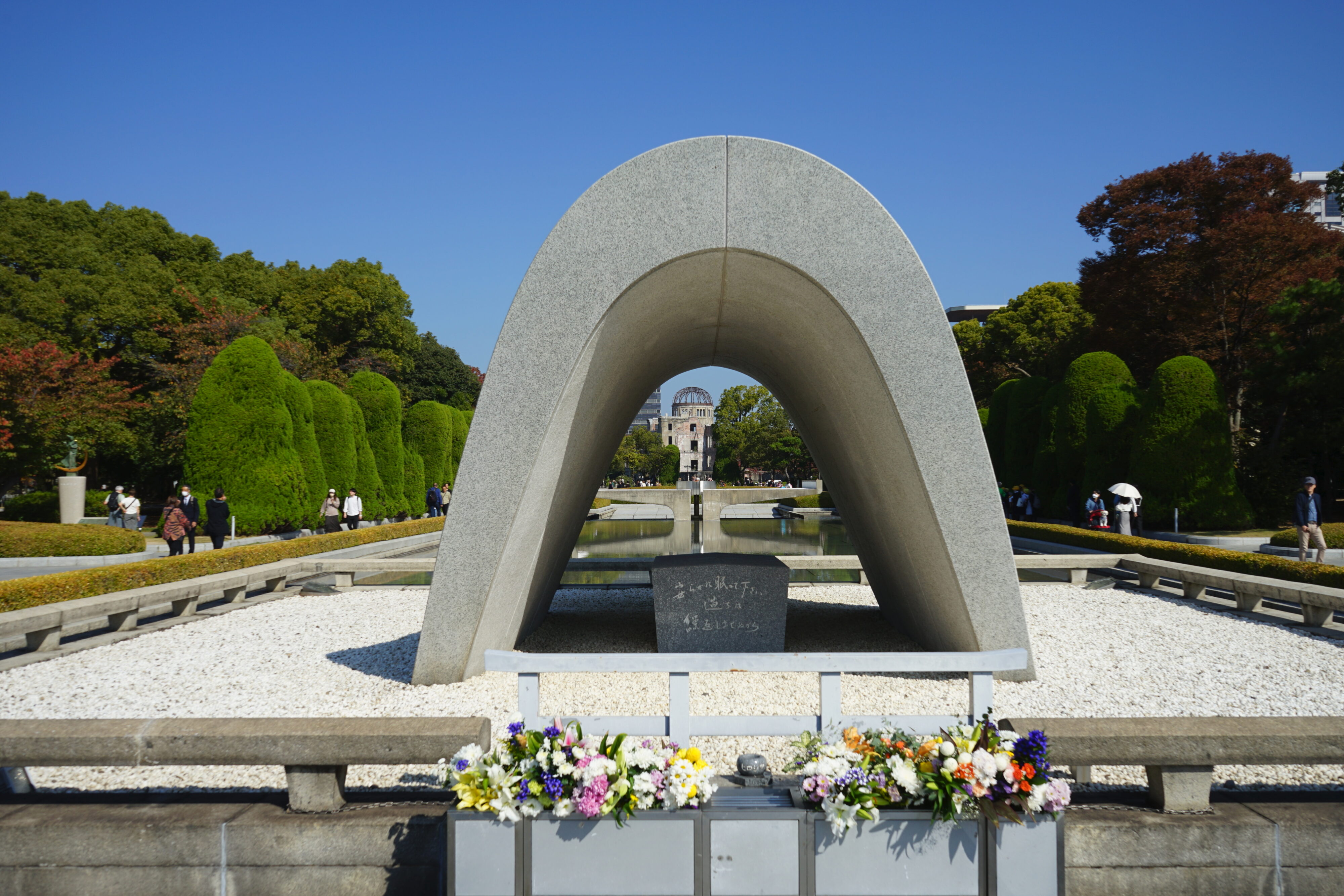 Blick auf das zentrale Erinnerungsmonument im Friedenspark: Ein Betonbogen steht auf eine mit Kieselsteinen bedeckten Fläche. Durch den Bogen lässt sich im Hintergrund die Atombombenkuppel erkennen.