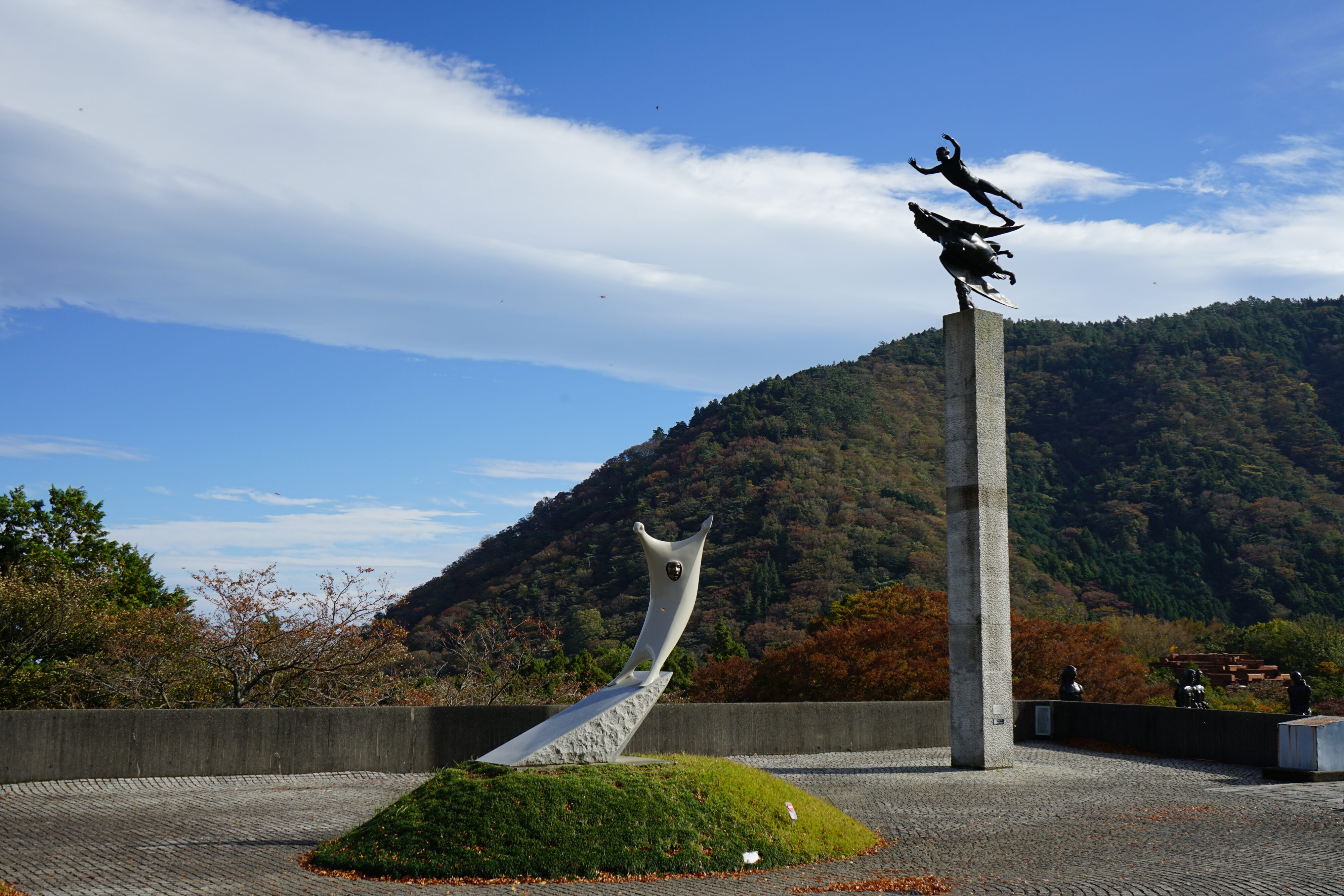 Verschiedene Skulpturen im Vordergrund und im Hintergrund ein grüner Hügel und blauer Himmel mit weißen Wolken.