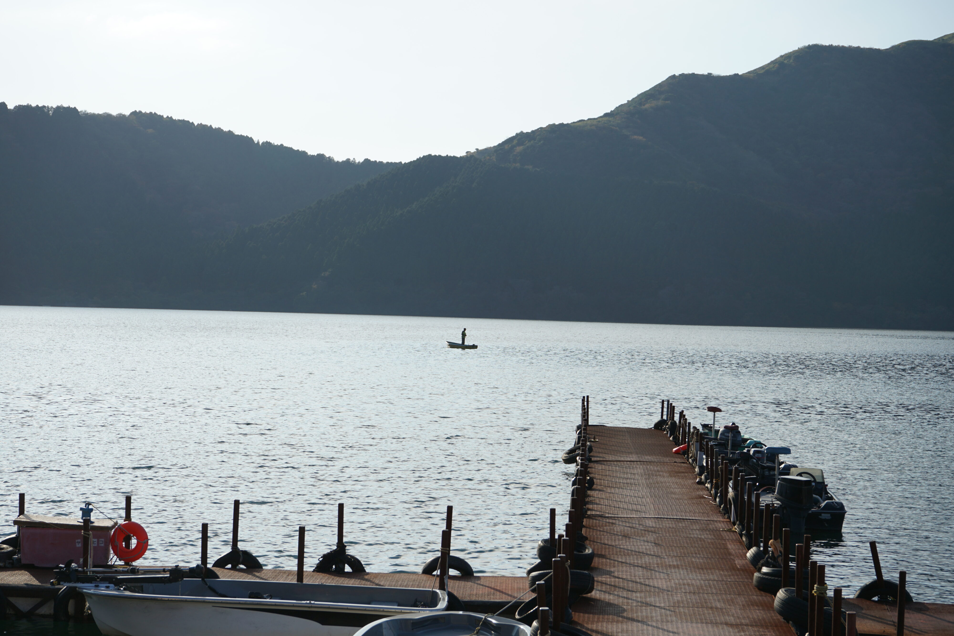 Blick auf den Ashi-See mit Steg im Vordergrund und Hügeln am anderen Ufer. Auf dem See ein Boot und eine darin stehende Person.