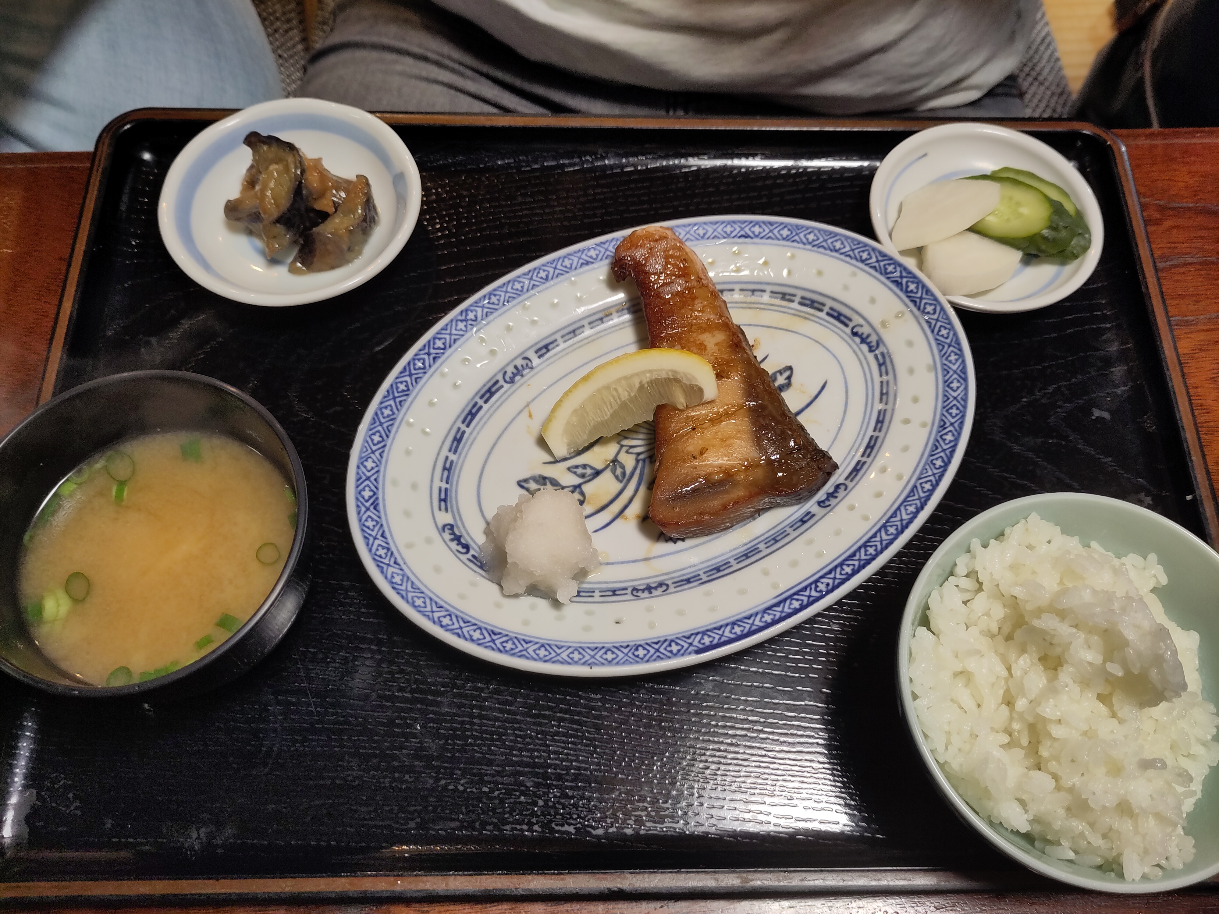 Blick auf das Fischmenü: mit einem Fischfilet in der Mitte, eine Schale Reis, eine Schale Misosuppe und zwei kleine Teller mit eingelegtem Gemüse und Fleisch. 