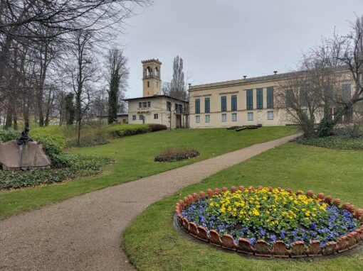 Blick auf die Rückseite des Schlosses Glienicke aus dem Garten heraus mit gepflegtem Rasen und runden Blumenbeeten.