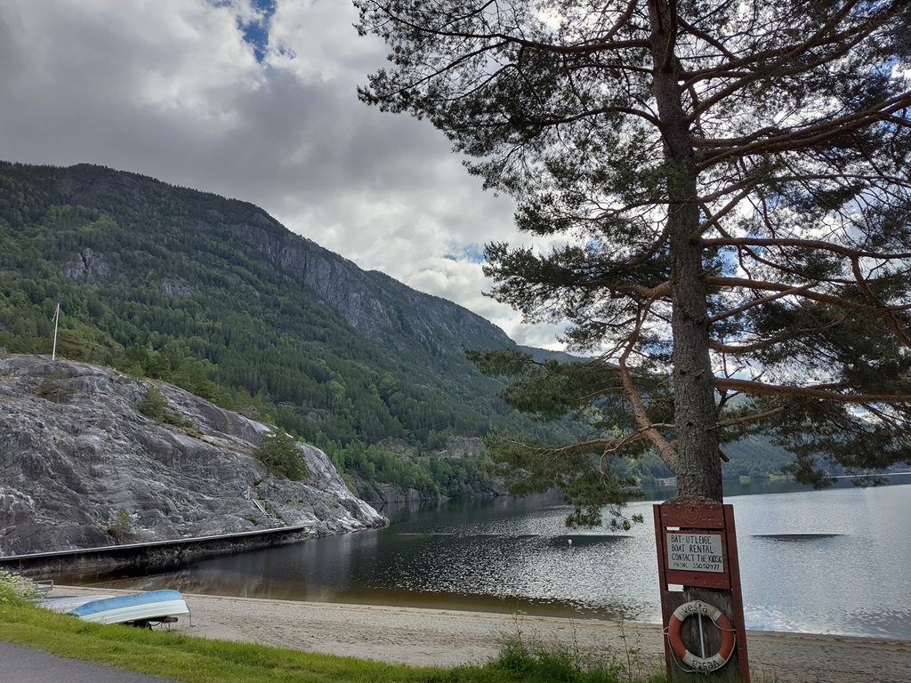 Blick auf das Ufer eines Sees. Im Vordergrund ein Strand mit einem umgedrehten Boot und ein Nadelbaum mit Rettungsring. Im Hintergrund Felswände von Bergen.