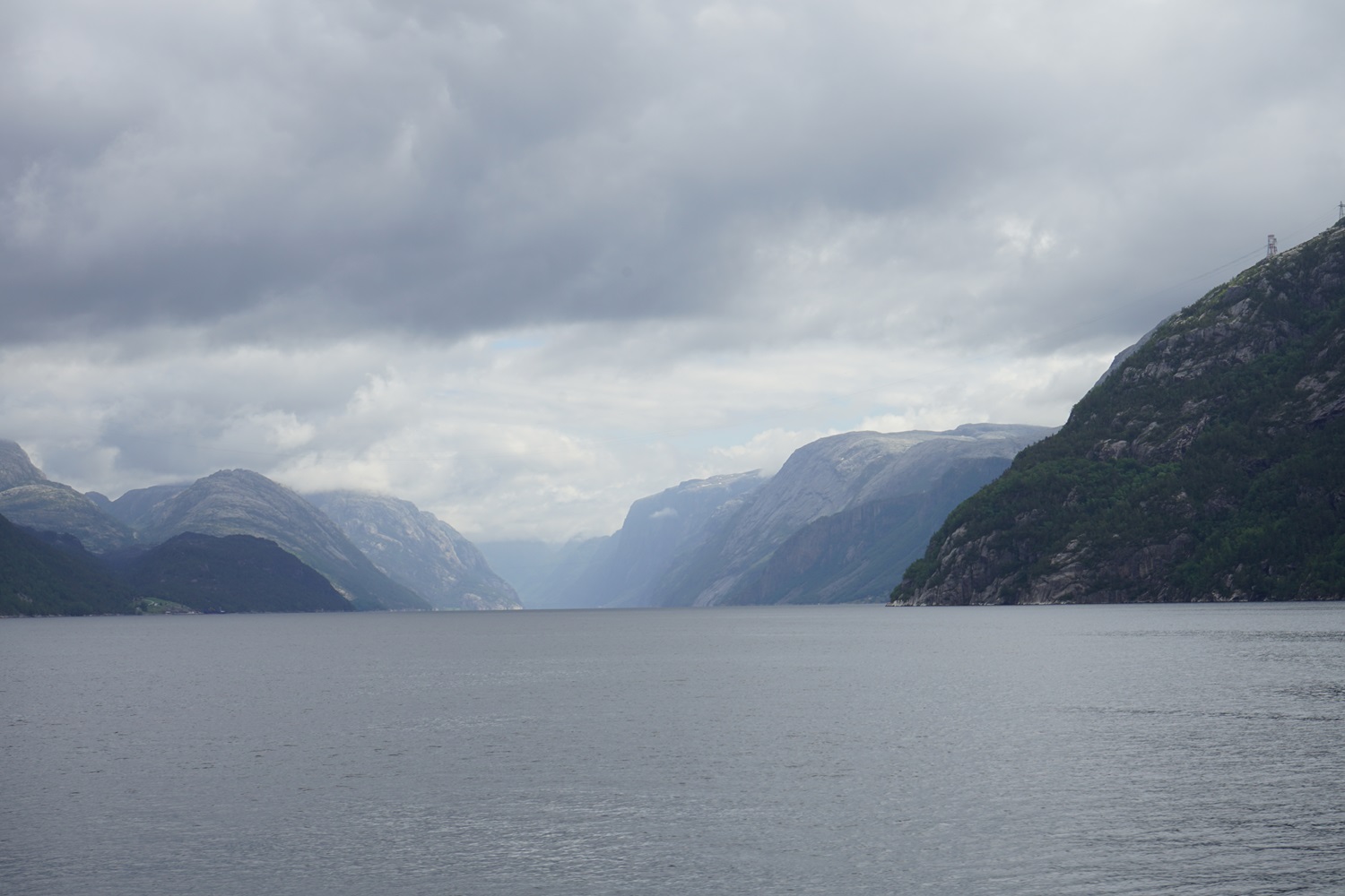 Blick in einem Fjord, der sich in der Mitte des Bildes immer weiter verjüngt umgeben von Bergen. Im Hintergrund graue Wolken.