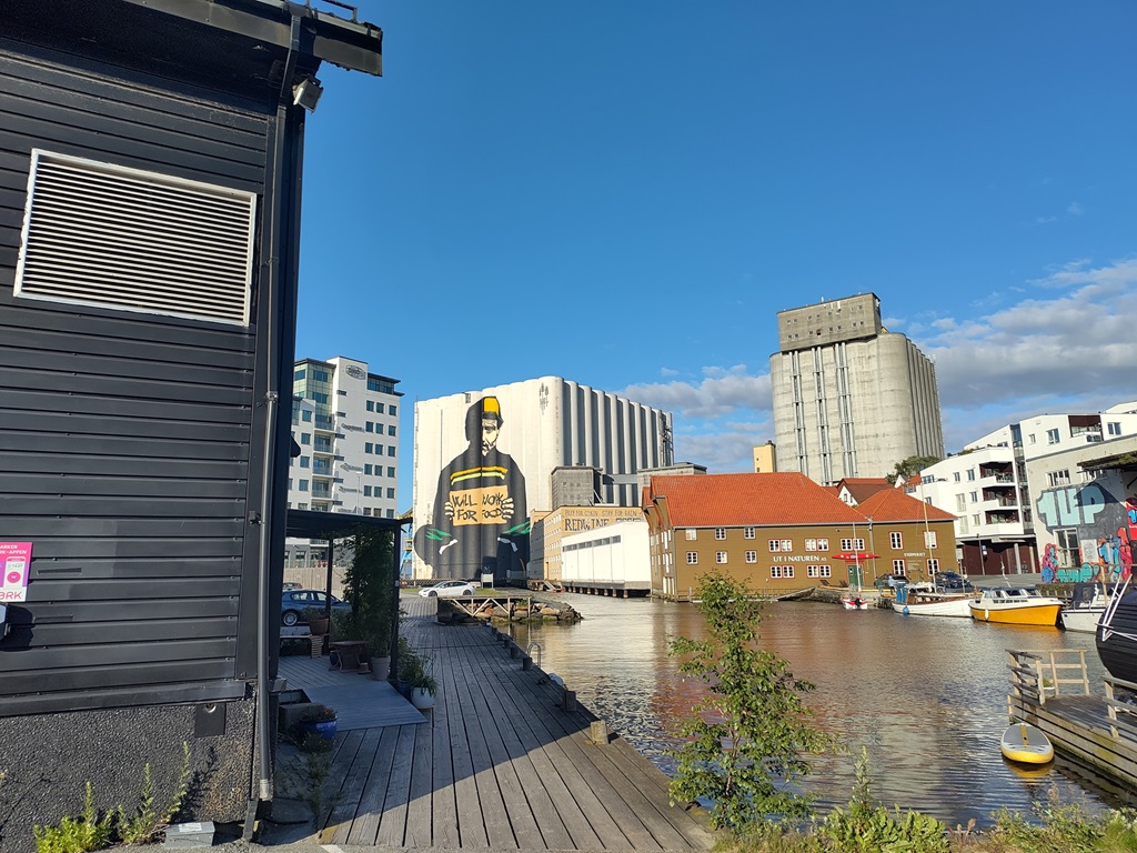 Blick auf einen Hafen mit Wohn- und Fabrikhäusern. An einem Gebäude befindet sich das Bild eines Mannes, der ein Schild hält auf dem steht "Will work for Food".