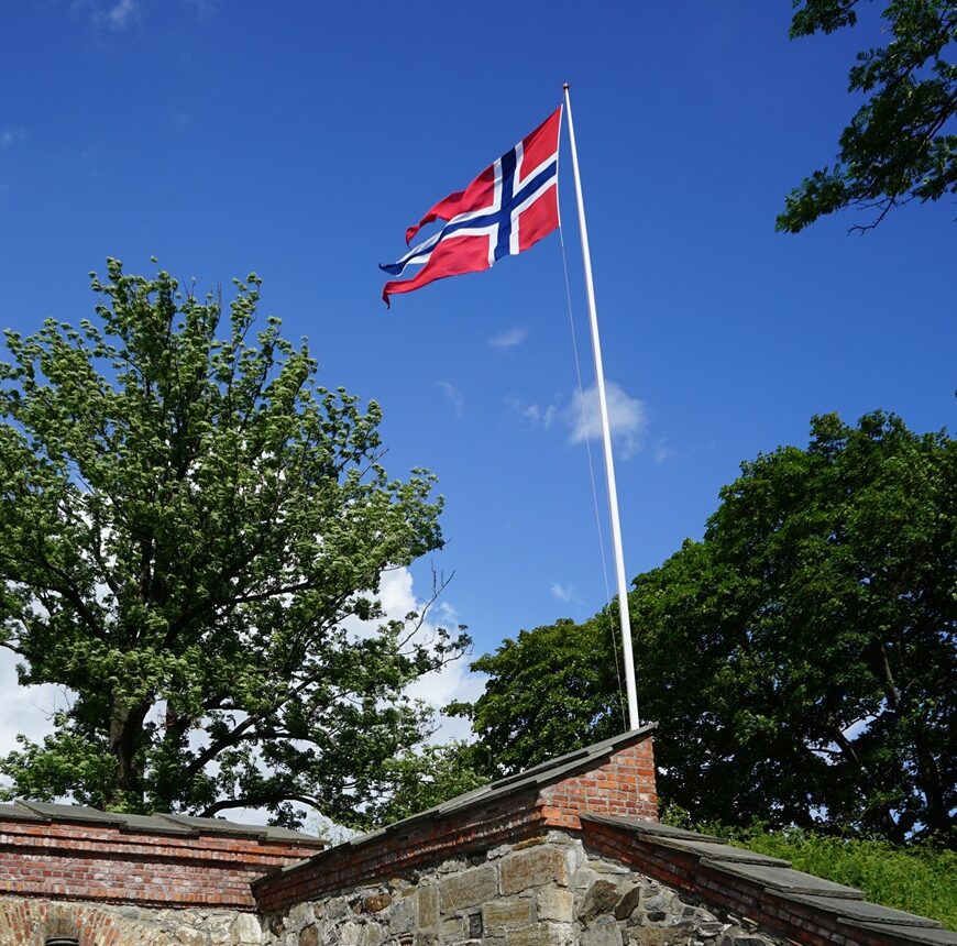 Norwegische Flagge auf einer Burgmauer. Im Hintergrund Bäume und blauer Himmel.