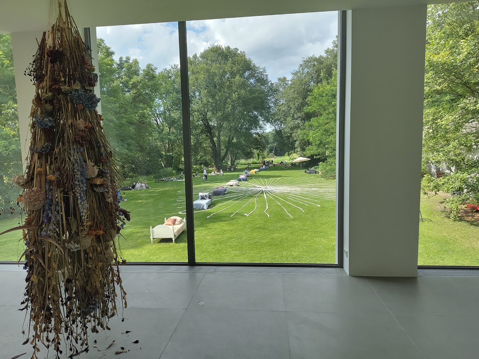 Blick auf den riesigen Trockenstrauß. Im Hintergrund eine Fensterfront mit Blick auf den Garten. Im Garten stehen Betten und sitzen Menschen.