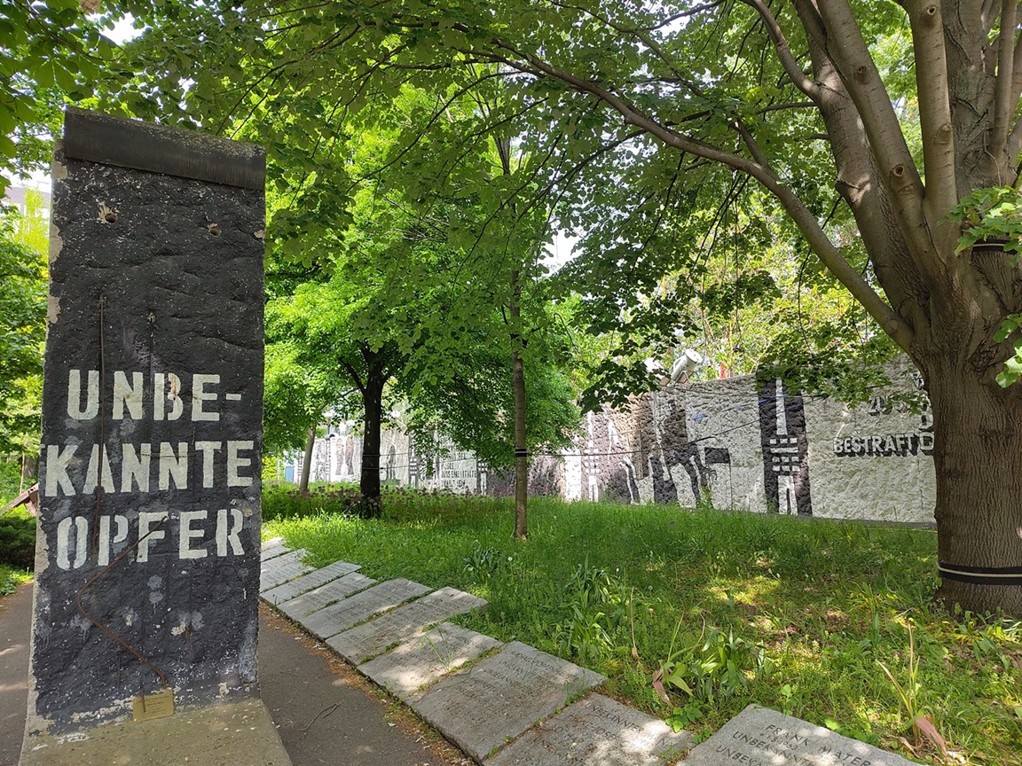 Mauerstück auf einem Weg auf dem "Unbekannte Opfer" steht. Am Wegesrand liegen Grabplatten. Ein Stück der bemalten Mauer und Bäume sind zu sehen.