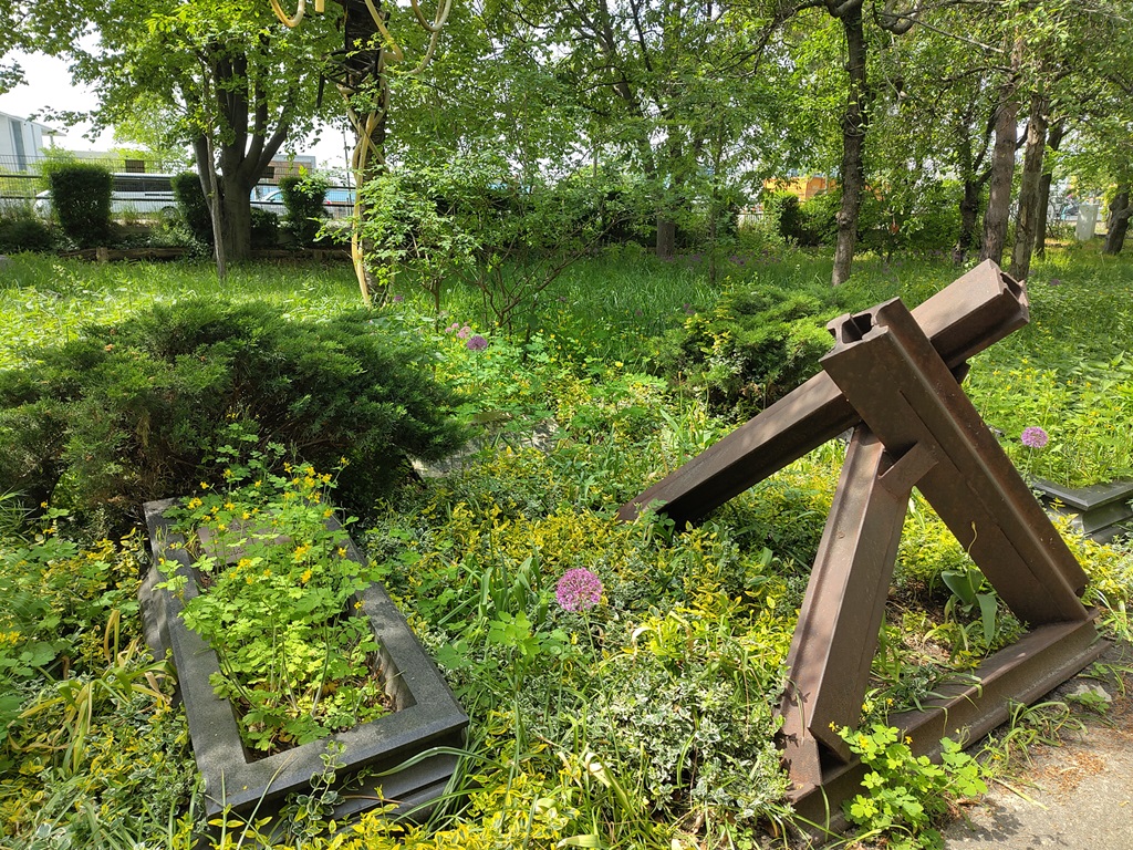 Panzersperre auf einer Wiese umgeben von Büschen und wuchernden Pflanzen, daneben ein Betonkasten, der an eine Grabumfassung erinnert.