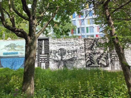 Blick auf Mauerreste mit dem Schriftzug "Parlament der Bäume" eingerahmt von Bäumen und Büschen.