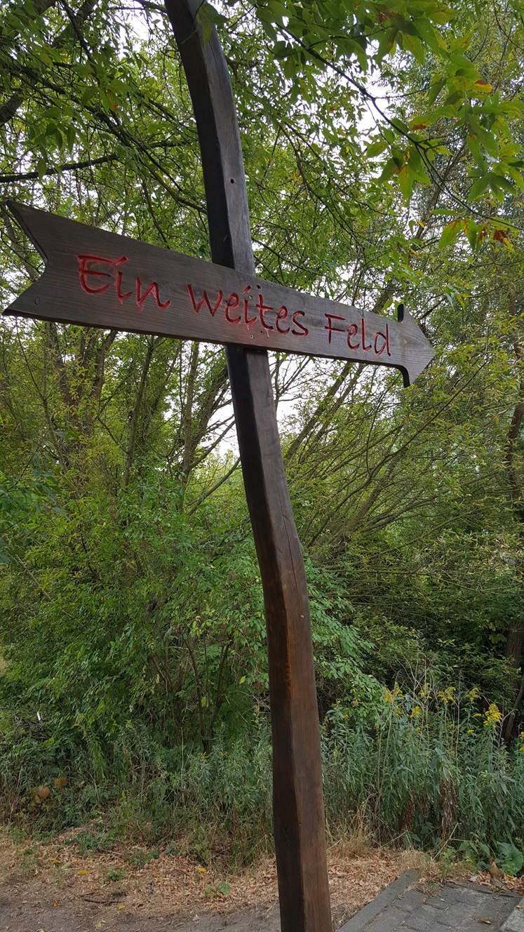 Im Vordergrund ein Holzschild auf dem "ein weites Feld" steht. Im Hintergrund grüne Bäume und Büsche.