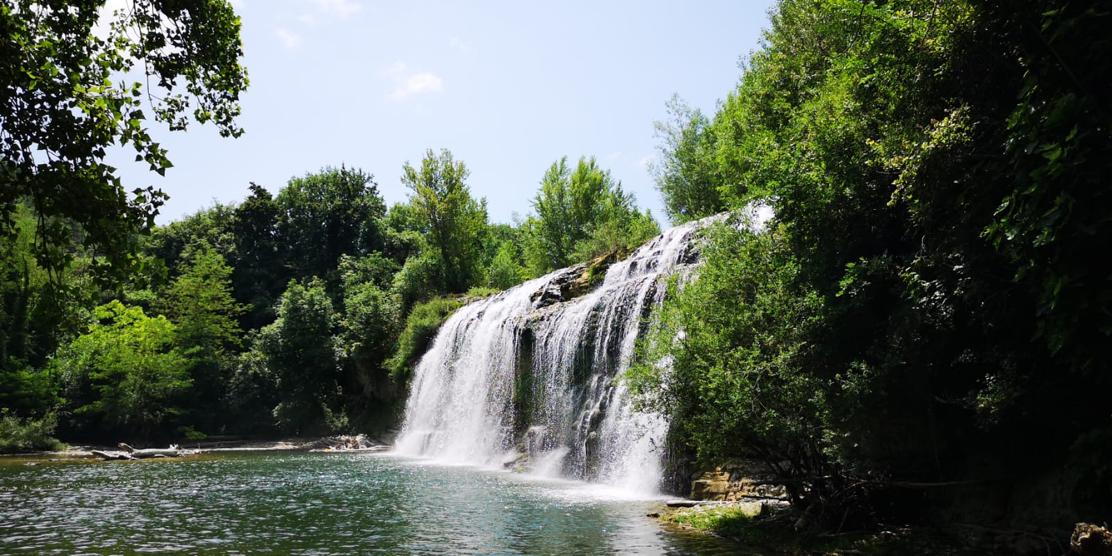 Kleiner so etwa 10 Meter hoher Wasserfall, umgeben von Bäumen und Büschen, der in einem kleinen See fällt. 