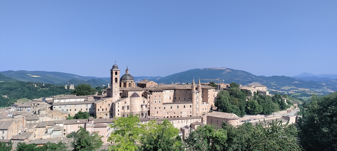 Blick auf Urbino vom Parco della Resistenza aus. Hinter den Gebäuden auf einem Hügel ragen weitere grüne Hügel am Horizont auf.