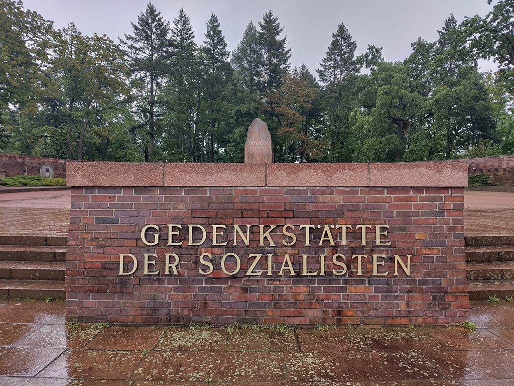 Ziegelmauer mit der Inschrift "Gedenkstätte der Sozialisten" im Hintergrund ein Gedenkstein, ein gemauertes Rondell und Bäume.