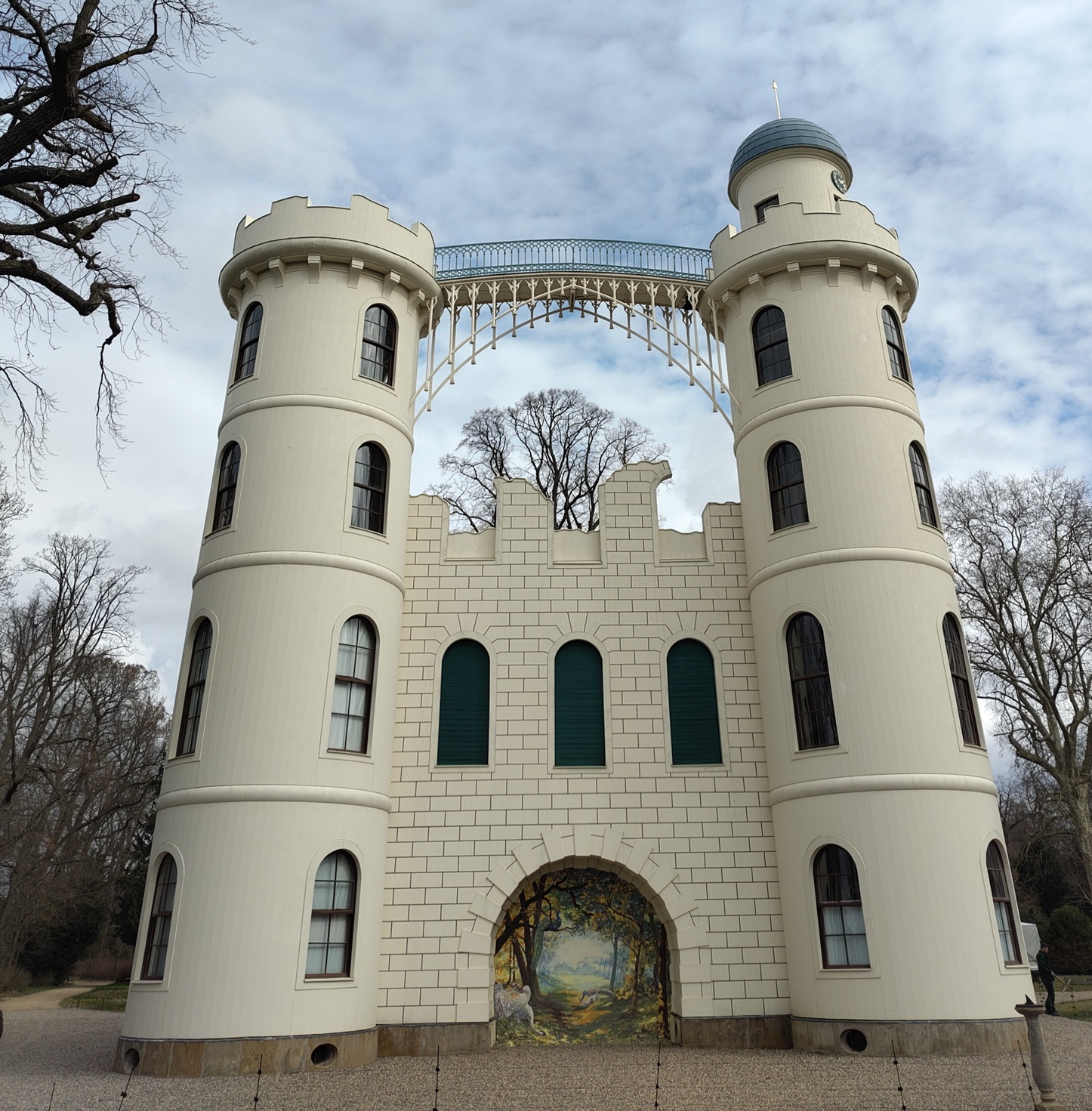 Seitenansicht des Schlosses auf der Pfaueninsel: Zwei Türmchen sind mit einer Brücke verbunden. Auf der Hauswand dazwischen ein Waldbild mit Pfau.