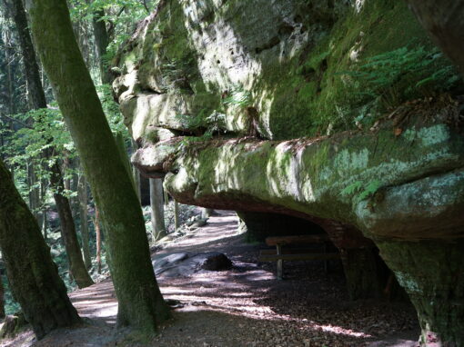 Felsen, der über einen Weg ragt. Unter dem Felsen ist ein Raststätte mit Bank und Tisch.