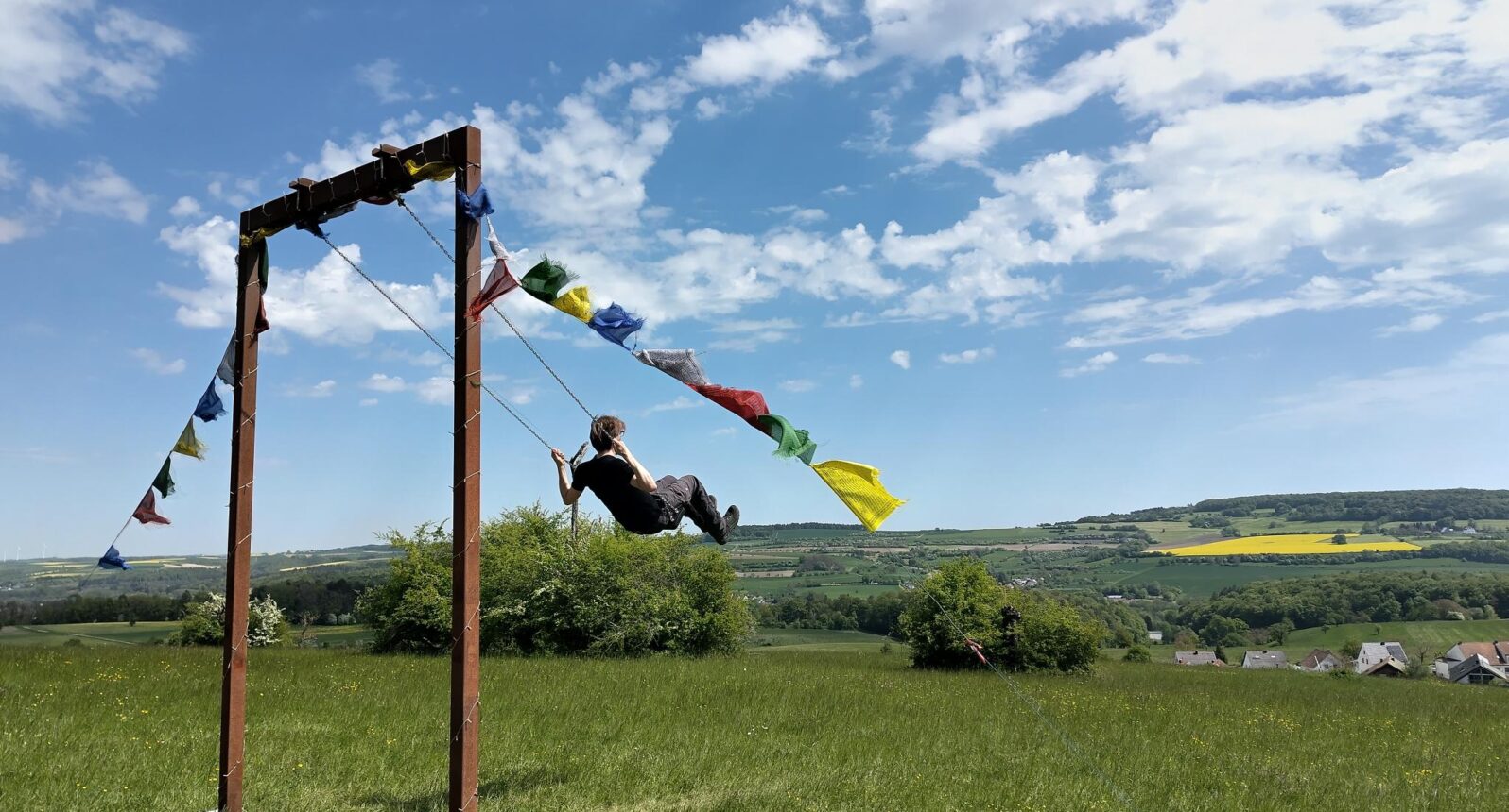 Schaukel auf einem Hügel, auf der ein Mann schaukelt. Im Hintergrund blauer Himmel und grüne Hügel.