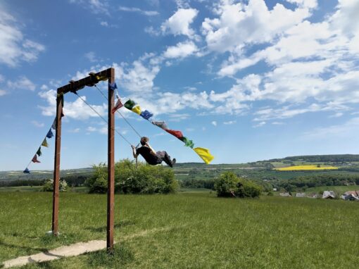 Schaukel auf einem Hügel, auf der ein Mann schaukelt. Im Hintergrund blauer Himmel und grüne Hügel.