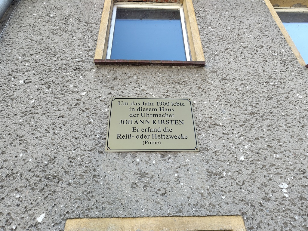Gedenktafel für Johann Kirsten, dem Erfinder der Heftzwecke, an einer grauen Hauswand.